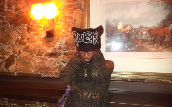 Aesthetic mage of me posed in a cat ear shaked beanie with the word "Queer" on it and red eyes from the camera flash. Wearing a knit green top that shows hits of my beautiful black skin